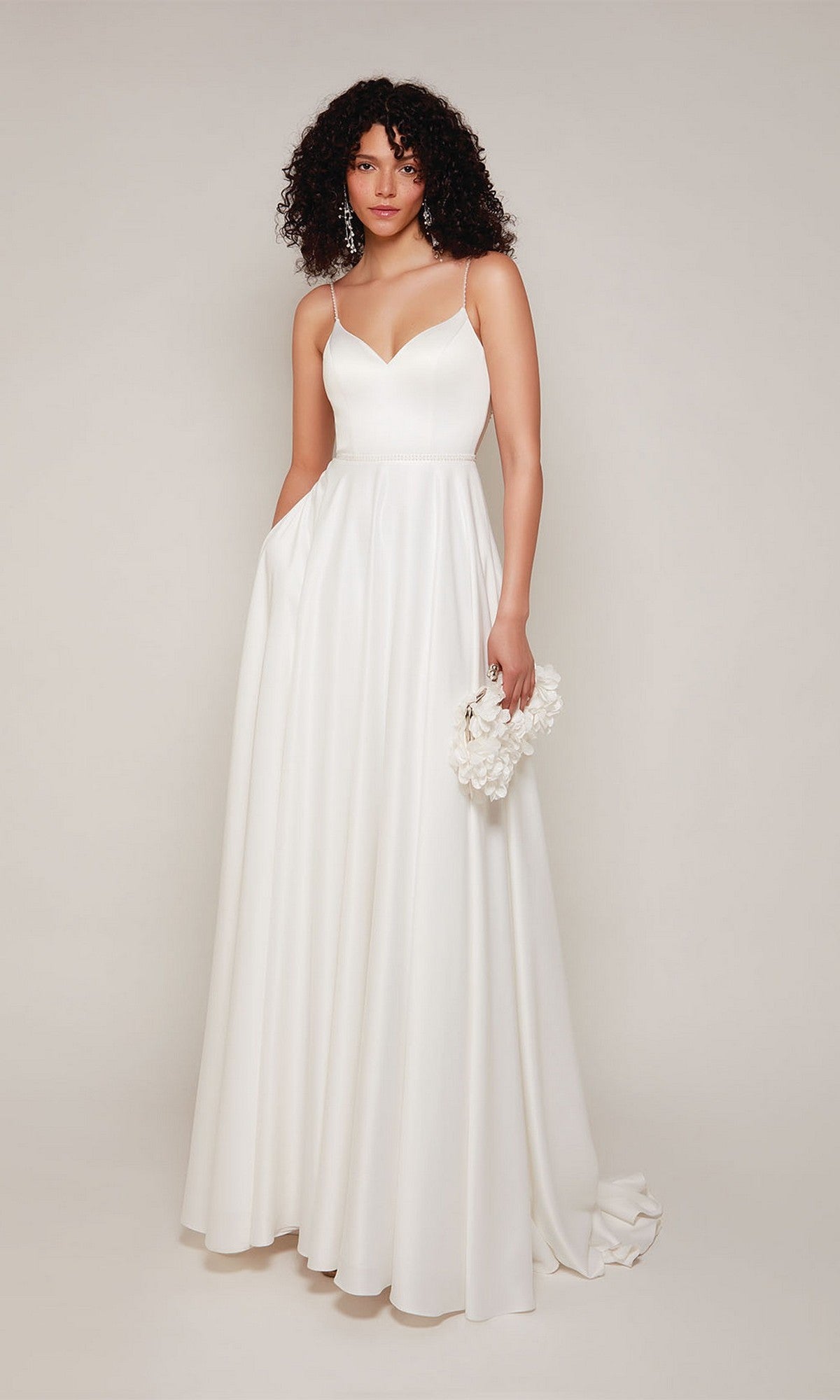 A woman in the Alyce 7099 long white dress with beaded spaghetti straps holds a small white floral purse while standing against a plain light background.
